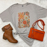 A styled flat-lay featuring a light gray short-sleeve t-shirt with a retro floral sun graphic in orange, brown, and cream. The t-shirt is styled with a knot at the hem and is accompanied by a bright orange crossbody bag and a pair of brown ankle boots on a white faux fur background.