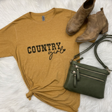 A styled flat-lay featuring a mustard yellow t-shirt with 'COUNTRY girl' printed in black on the front, tied at the bottom hem. The shirt is styled with a pair of brown leather ankle boots and an olive green crossbody purse on a white faux fur background.