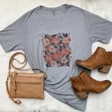 A styled flat-lay featuring a heather gray short-sleeve t-shirt with a large coral and blush floral graphic on the front, accompanied by a pale pink crossbody bag and brown ankle boots on a white faux fur background.