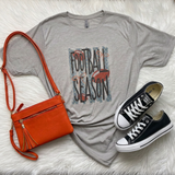 A styled flat-lay outfit featuring a light gray graphic t-shirt with 'FOOTBALL SEASON' text, an orange crossbody bag, and black canvas sneakers, all arranged on a white faux fur background.