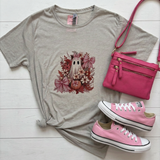 A styled flat-lay featuring a light gray t-shirt with a cute ghost and autumn floral graphic, accompanied by a hot pink crossbody bag, and pink canvas sneakers  on a white wood background.