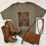 A styled flat-lay featuring an olive green graphic t-shirt with a colorful 'Just a Girl who loves Country Music' print, accompanied by a brown leather crossbody bag and a pair of brown western boots on a white faux fur background.
