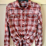A long-sleeve button-up flannel shirt in a rust red, brown, and white plaid pattern, styled with the bottom tied in a knot, hanging on a dark hanger against a light beige background.