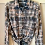 A long-sleeve button-up flannel shirt with a distressed bleach-splatter plaid pattern in navy blue, gray, and beige, featuring two chest pockets and a tied waist.