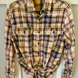 A long-sleeve plaid button-up shirt in tan, brown, and black, tied at the waist and photographed against a light-colored background.