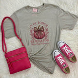 A styled flat-lay featuring a light gray graphic t-shirt with a pink pumpkin design and the text 'IN OCTOBER WE WEAR PINK', paired with a hot pink crossbody bag and pink low-top sneakers on a white faux fur background.